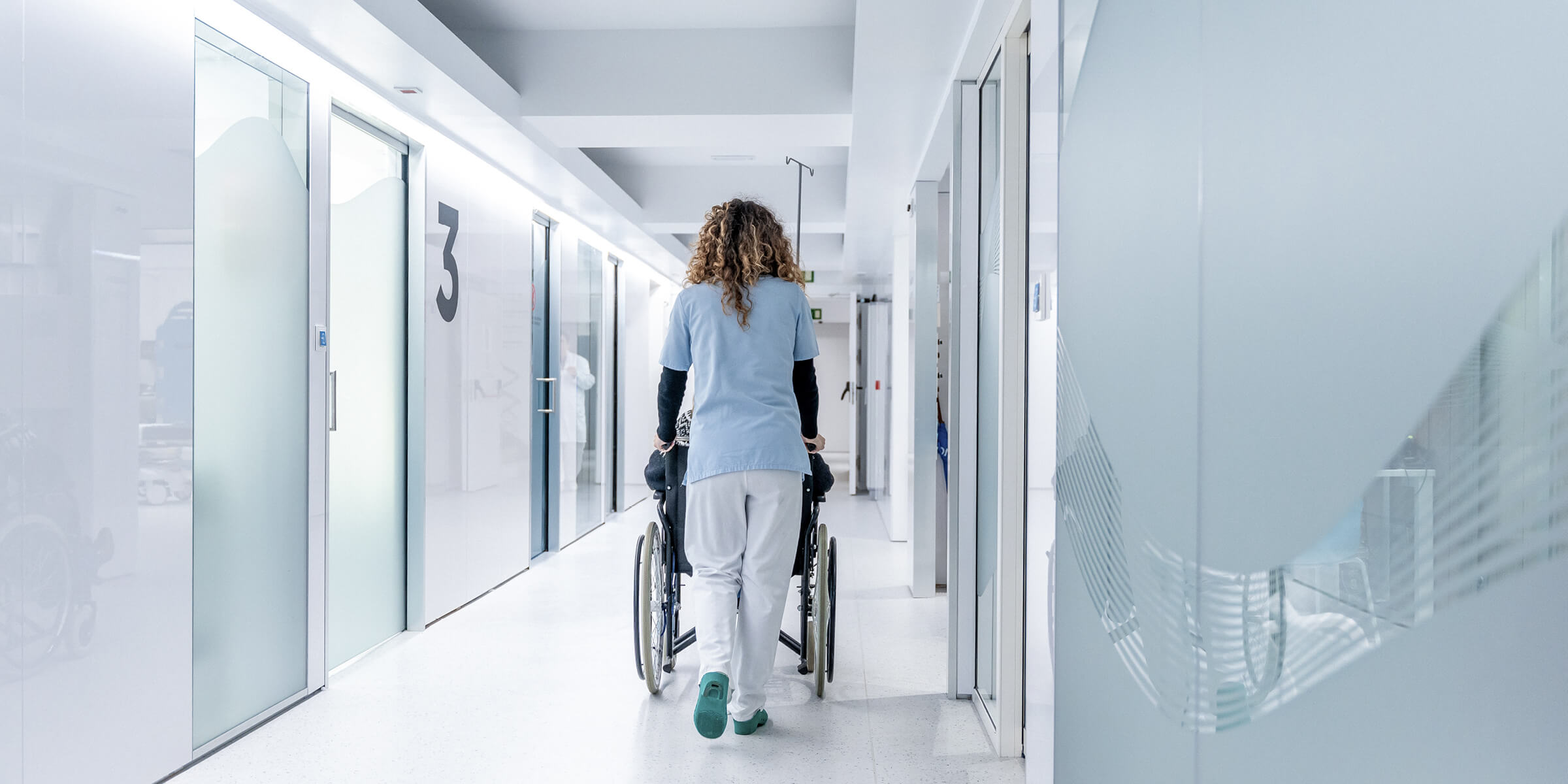 Bild zeigt die Rückansicht einer Krankenschwester, die einen Rollstuhl durch das Krankenhaus schiebt
